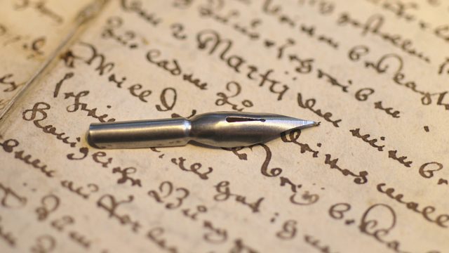 Una hoja antigua escrita con una pluma. Encima de esa hoja está la punta de esa pluma