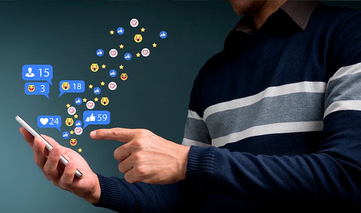 Una mano tocando un móvil de donde salen iconos típicos de redes sociales como los Me gusta, los corazones y demás.