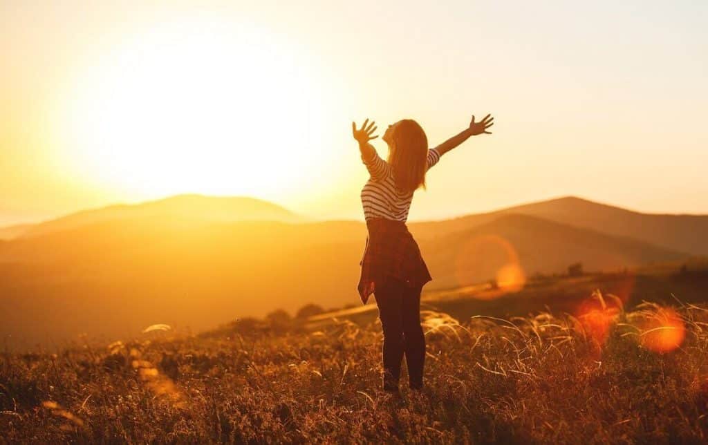 Una chica de pie y abriendo los brazos de forma feliz mientras sale el amanecer al fondo